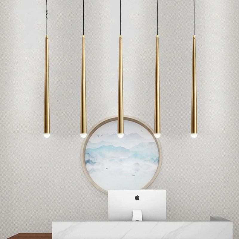 Five slim brass tube chandelier lights hang above a marble counter with a computer monitor and round wall art.