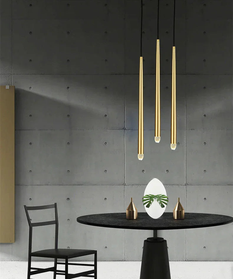 Brass chandelier with three tapered tubes and crystal tips hangs above a round black dining table against a gray concrete wall.