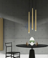 Brass chandelier with three tapered tubes and crystal tips hangs above a round black dining table against a gray concrete wall.