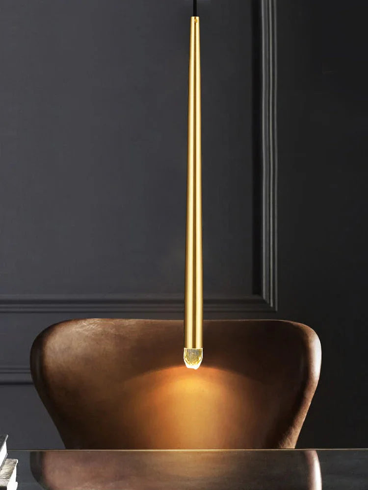 Single slim brass tube chandelier pendant glows above a brown leather chair against dark gray paneled walls.