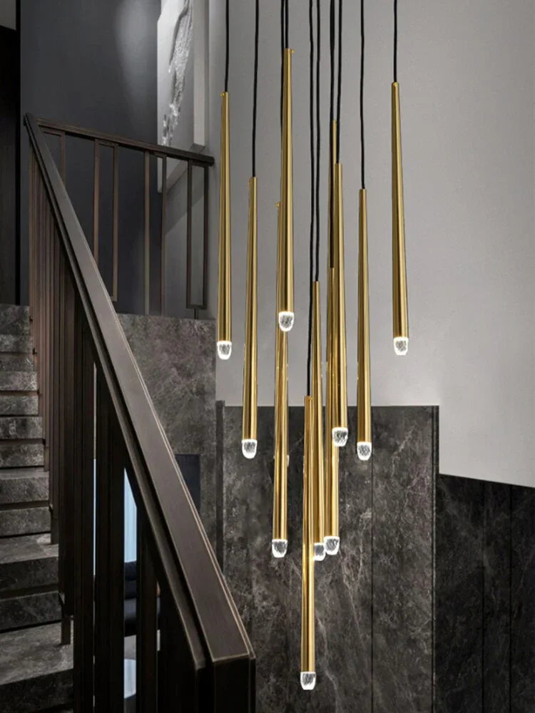 Brass chandelier with crystal-tipped tubes cascades at staggered heights beside a dark stone staircase and black railing.