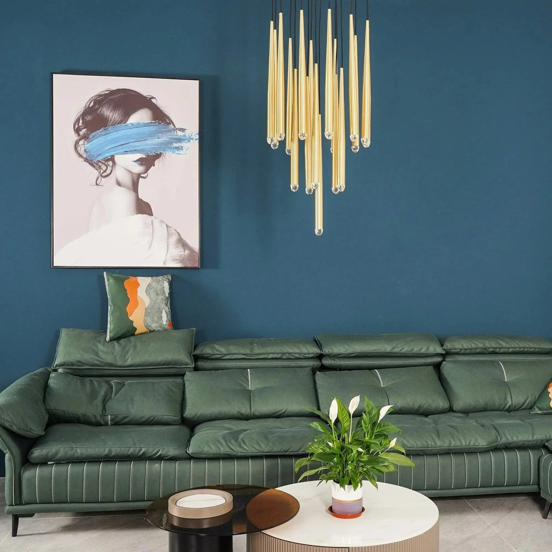 Brass chandelier with clustered crystal-tipped tubes hangs above a green sofa against a blue wall beside framed portrait artwork.