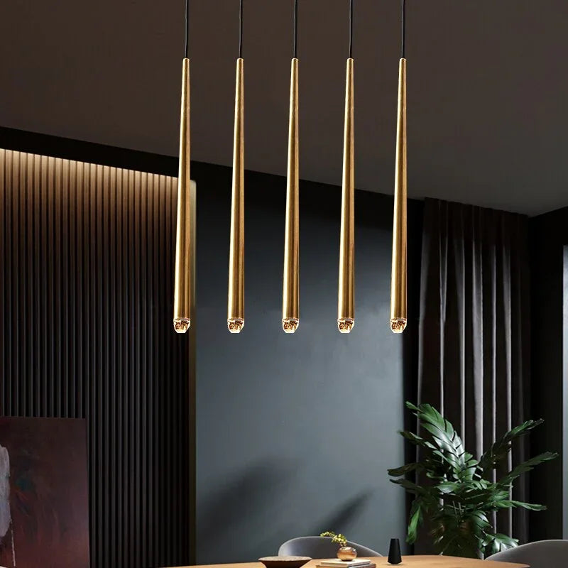 Brass chandelier with five crystal-tipped tubes hangs in a straight row above a dining table beside dark slatted walls.