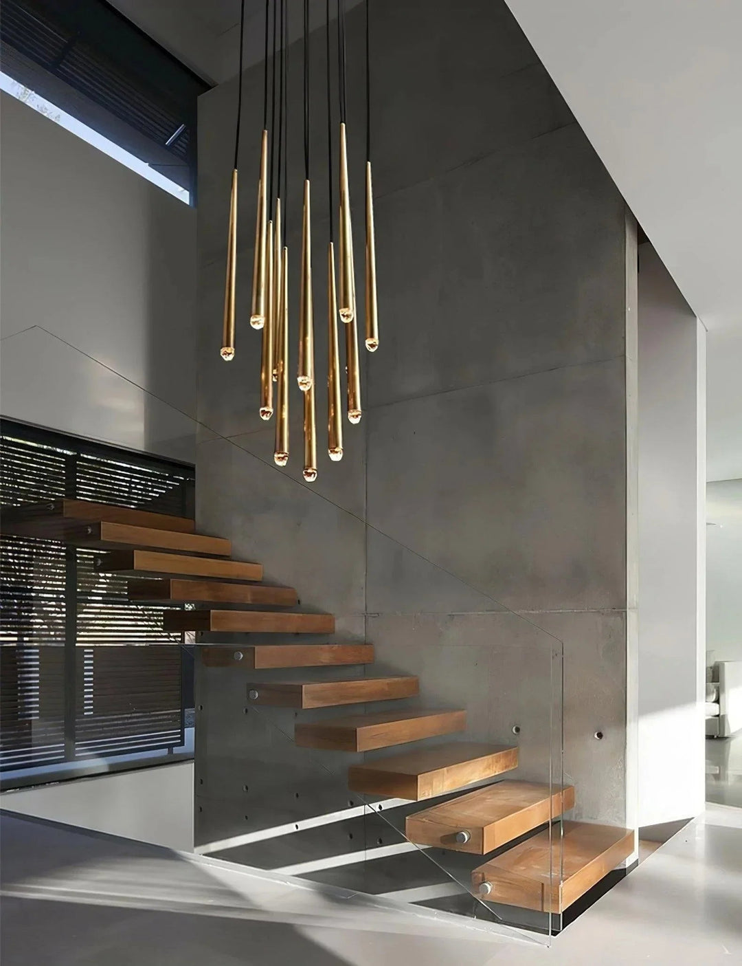Cluster of slim brass tube chandelier lights hangs above floating wooden staircase steps against a gray concrete wall.