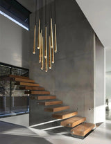 Cluster of slim brass tube chandelier lights hangs above floating wooden staircase steps against a gray concrete wall.