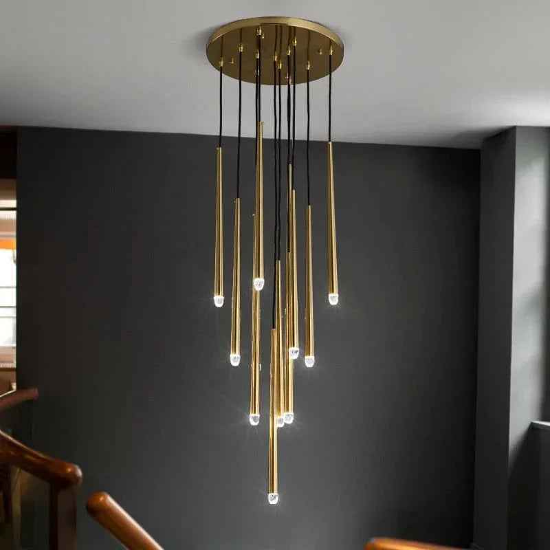 A modern chandelier with a circular gold ceiling mount, multiple vertical gold rods, black hanging cords, and small glowing bulbs, shown installed in a real interior with a dark gray wall and staircase railings.
