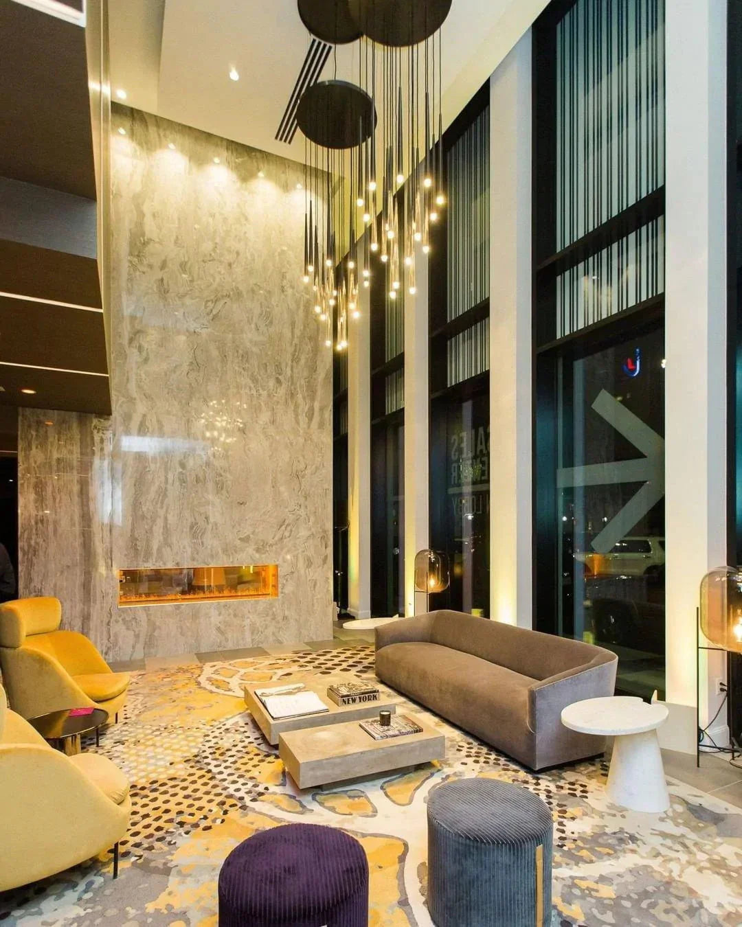 Illuminated brass tube chandelier hangs from circular ceiling plates in a double-height living room with tall windows and a fireplace.