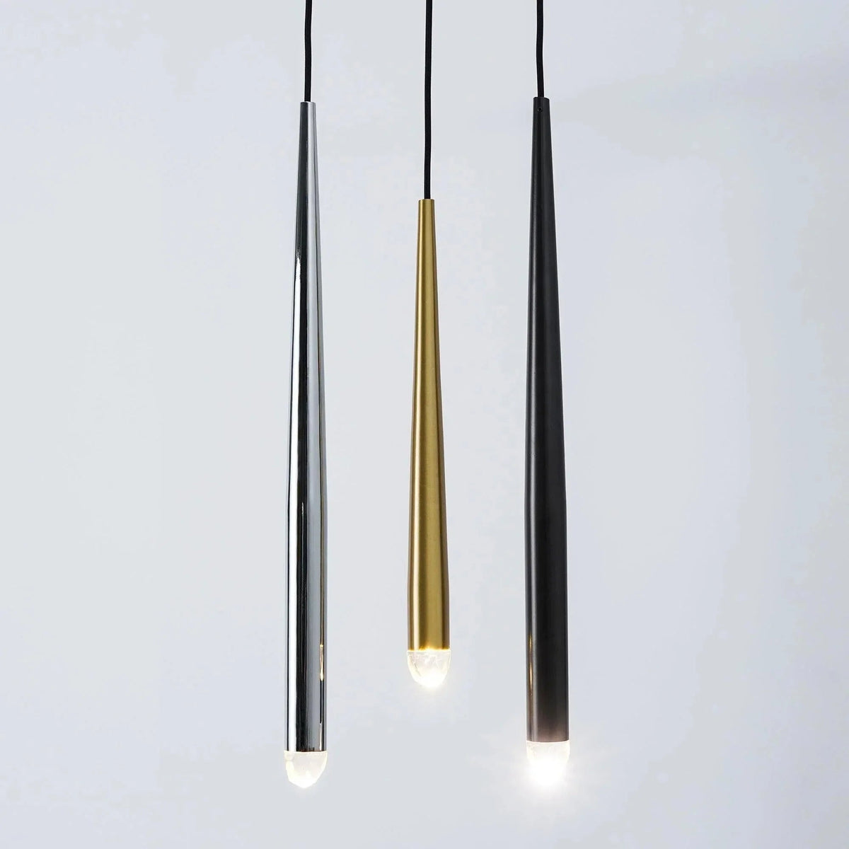 Three tapered chandelier tubes in silver, gold, and black finishes hang against a pale gray background with illuminated crystal tips.