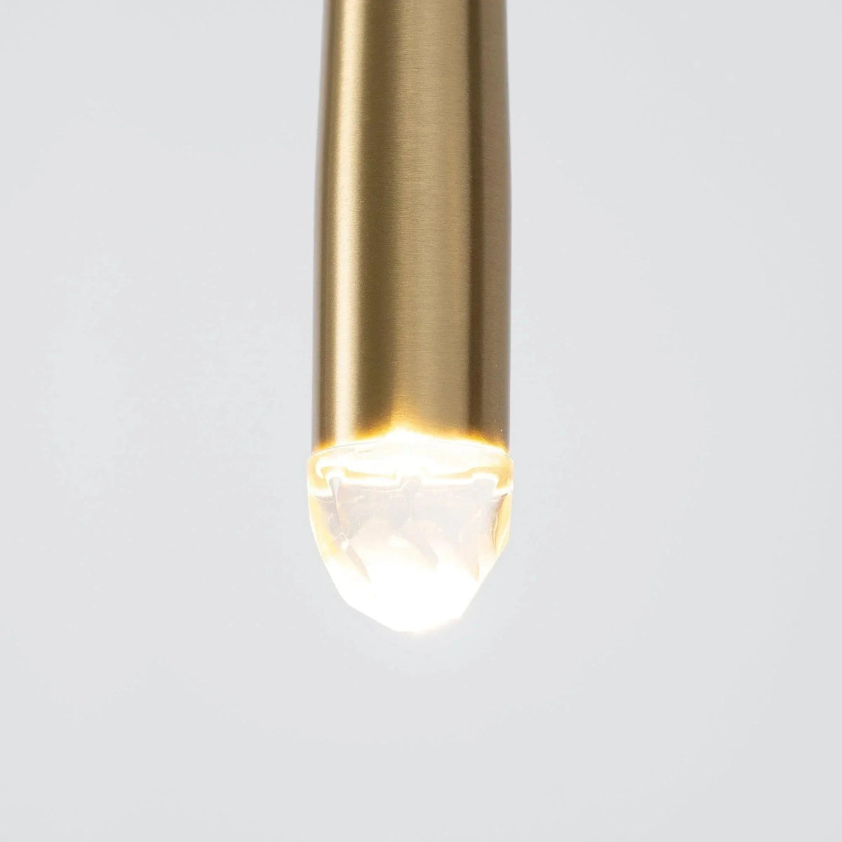 Close-up of a chandelier brass tube with an illuminated crystal tip against a pale gray background.