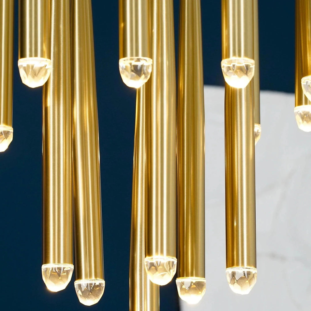 Brass chandelier tubes hang at staggered heights with glowing crystal tips against a dark blue and pale gray background.