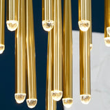 Brass chandelier tubes hang at staggered heights with glowing crystal tips against a dark blue and pale gray background.