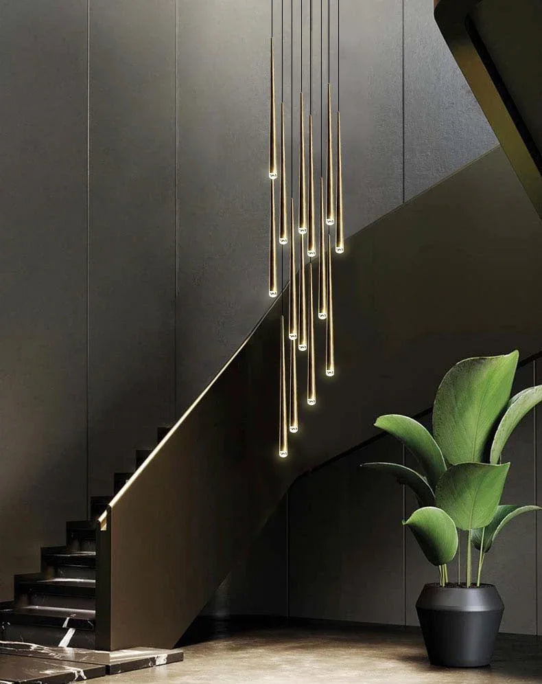 A modern multi-light linear pendant chandelier with warm glowing vertical bulbs hangs over a dark staircase in a contemporary interior setting with a potted green plant in the foreground.