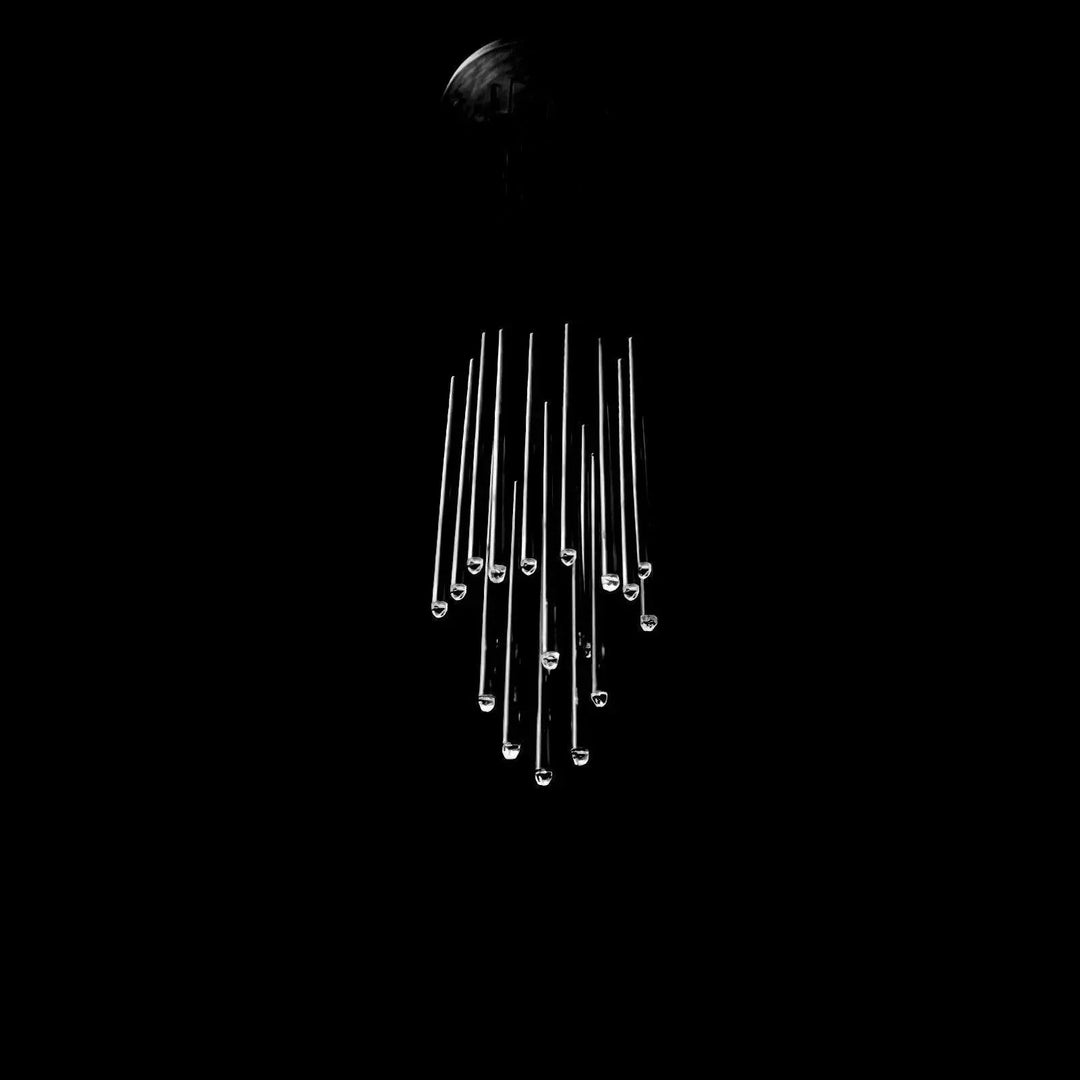 A modern chandelier with multiple clear glass rods of varying lengths hanging from a dark ceiling mount against a solid black background.
