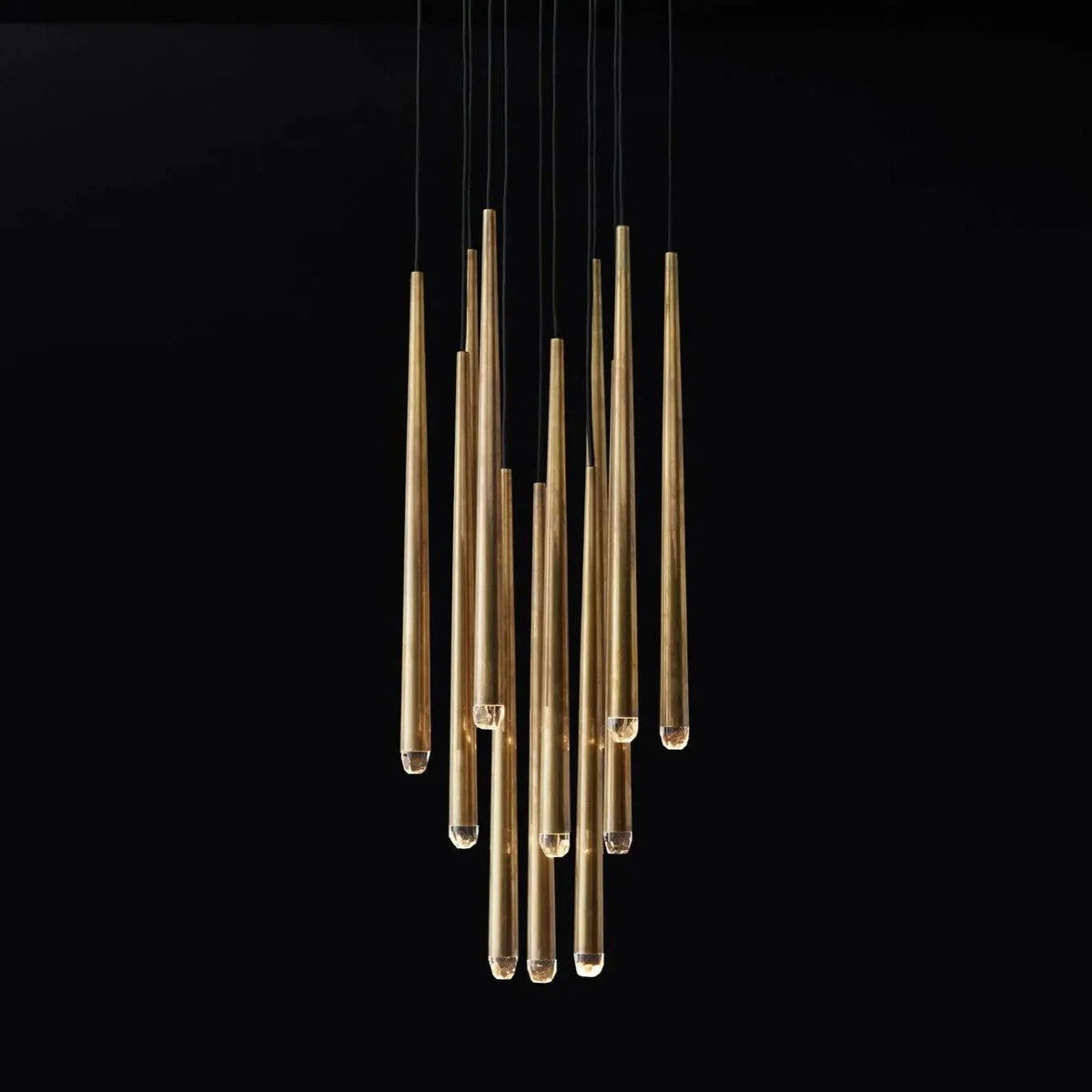 A modern multi-light linear chandelier with brushed gold metal tubes suspended by thin black cords against a solid black background.
