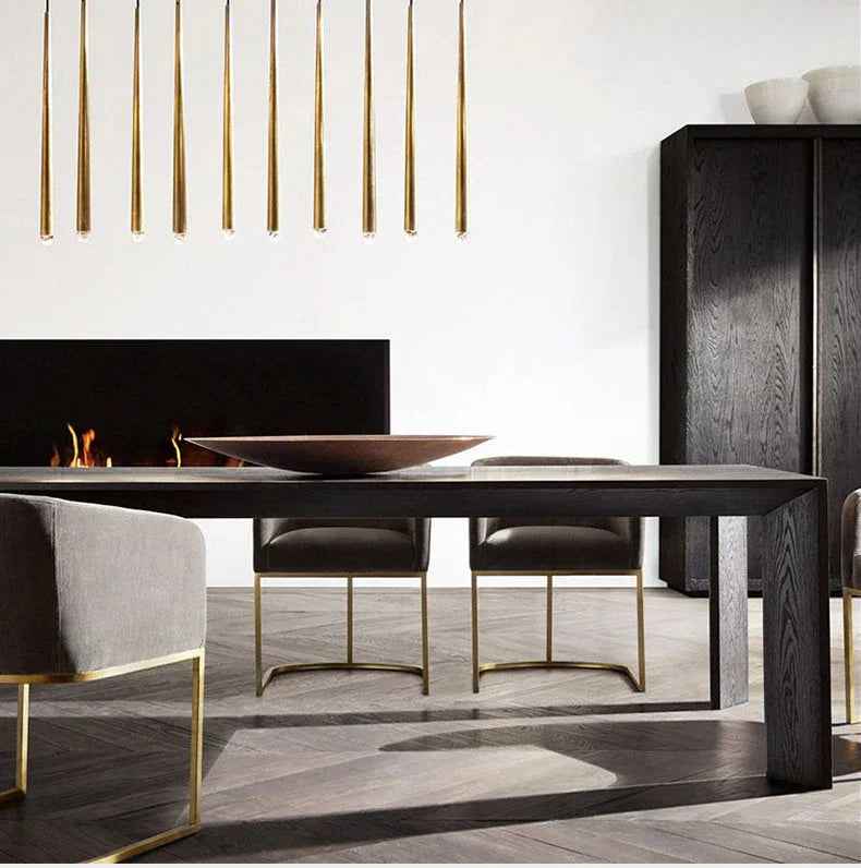 Slim brass tube chandelier drops in a row above a dark wood dining table beside a black fireplace and cabinet.