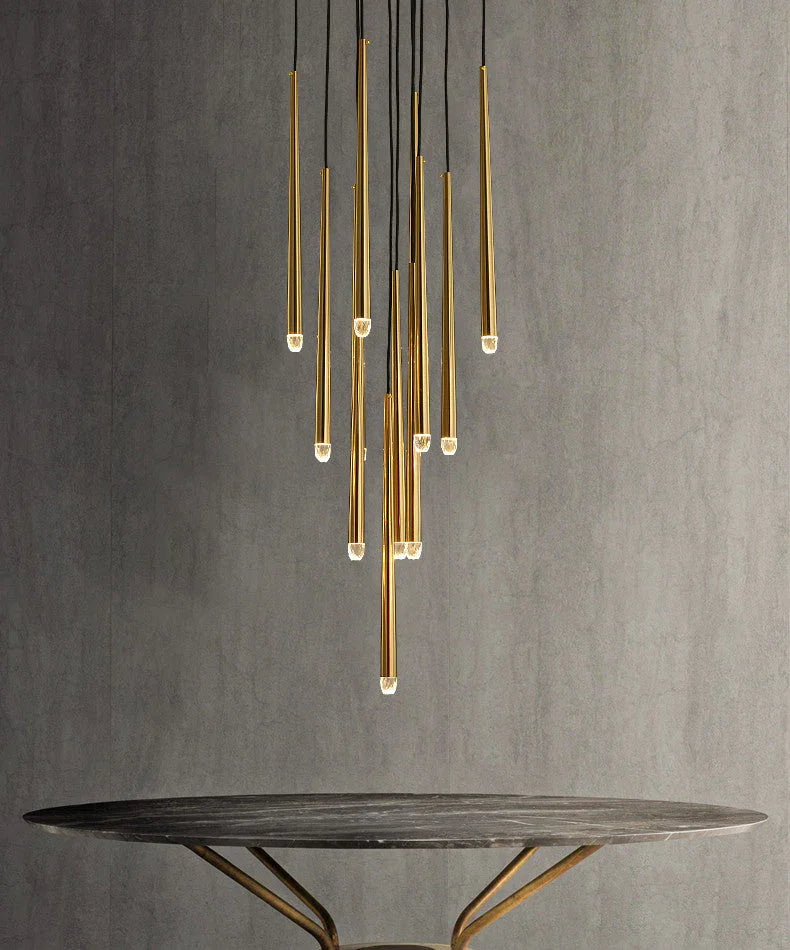 A modern multi-light chandelier with vertical brushed gold metal tubes suspended from black cords, shown above a round dark table against a textured gray wall.
