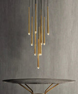 A modern multi-light chandelier with vertical brushed gold metal tubes suspended from black cords, shown above a round dark table against a textured gray wall.