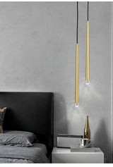 Two slim brass tube chandelier pendants hang beside a bed with gray bedding and a white nightstand.