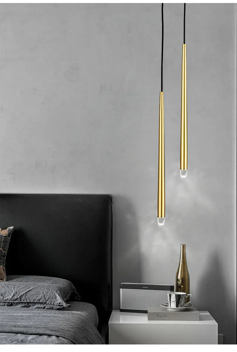 Two slim brass tube chandelier pendants hang beside a bed with gray bedding and a white nightstand.