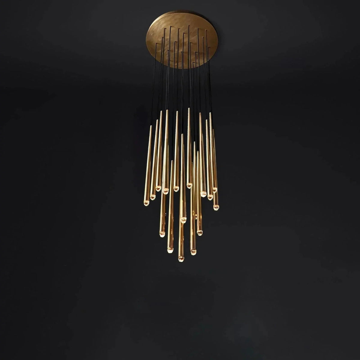 A modern chandelier with a circular gold ceiling mount and multiple hanging gold glass rods suspended by black cords, shown against a dark black background.
