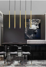 Row of five slim brass tube chandelier lights above a dark dining table with black chairs and marble floor.