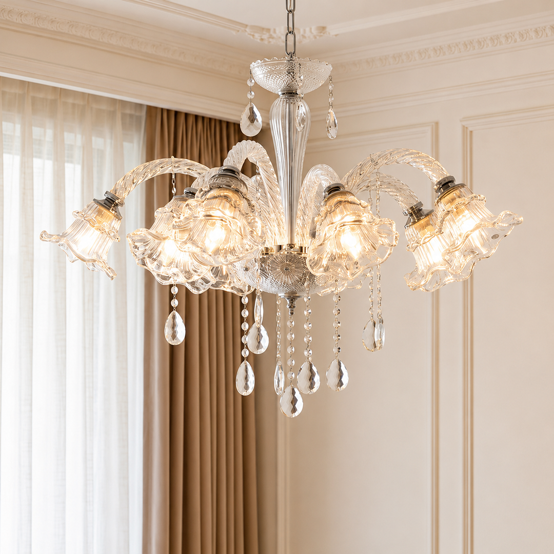 An ornate six-light crystal chandelier hanging in a classic cream-colored room with beige curtains and white paneling. The chandelier features clear glass arms and teardrop crystal pendants, with warm white bulbs glowing inside.