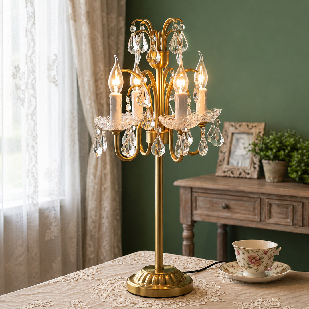 A gold brass table lamp with multiple candle-style lights and hanging clear crystal prisms, displayed on a table in a styled room with a green wall, wooden cabinet, curtain, and a floral teacup.
