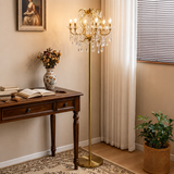 A gold-toned crystal chandelier floor lamp stands in a warmly lit room beside a wooden desk, curtains, and a potted plant.