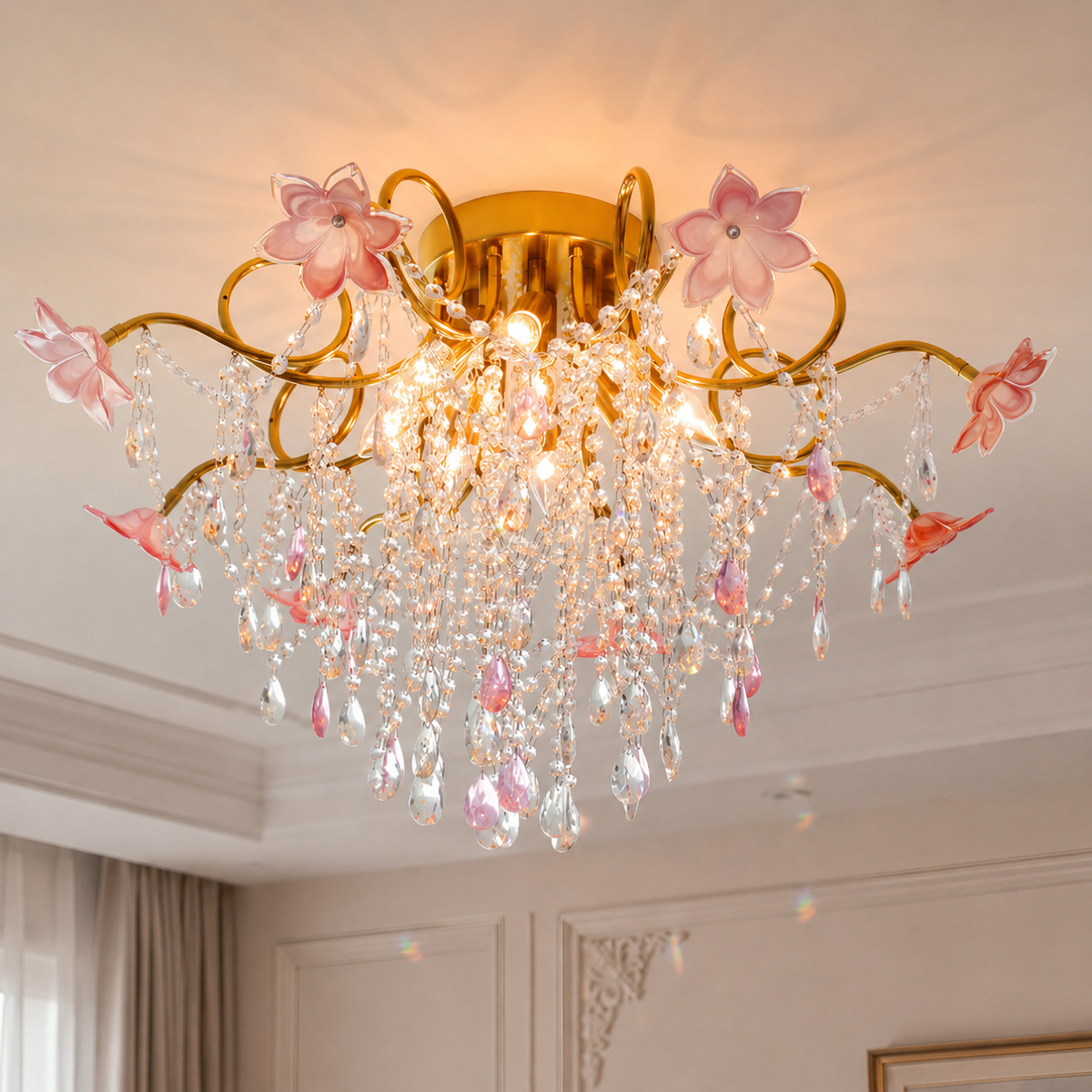 Illuminated chandelier with gold-tone branches, pink glass flowers and crystal droplets beneath a white ceiling in a room with beige curtains.