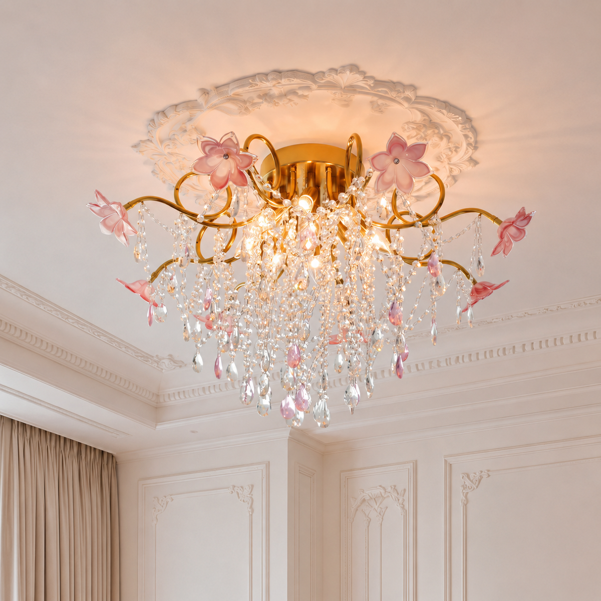 Illuminated chandelier with gold-tone branches, pink glass flowers and hanging crystals beneath an ornate white ceiling medallion.