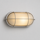 A silver wall sconce with ribbed frosted glass glows warmly, mounted horizontally on a light gray wall.