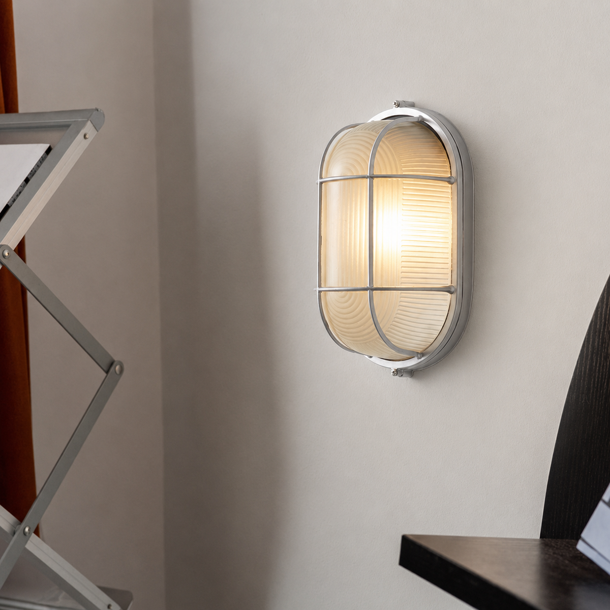 A silver cage wall sconce with ribbed frosted glass glows warmly on a pale wall beside dark furniture.