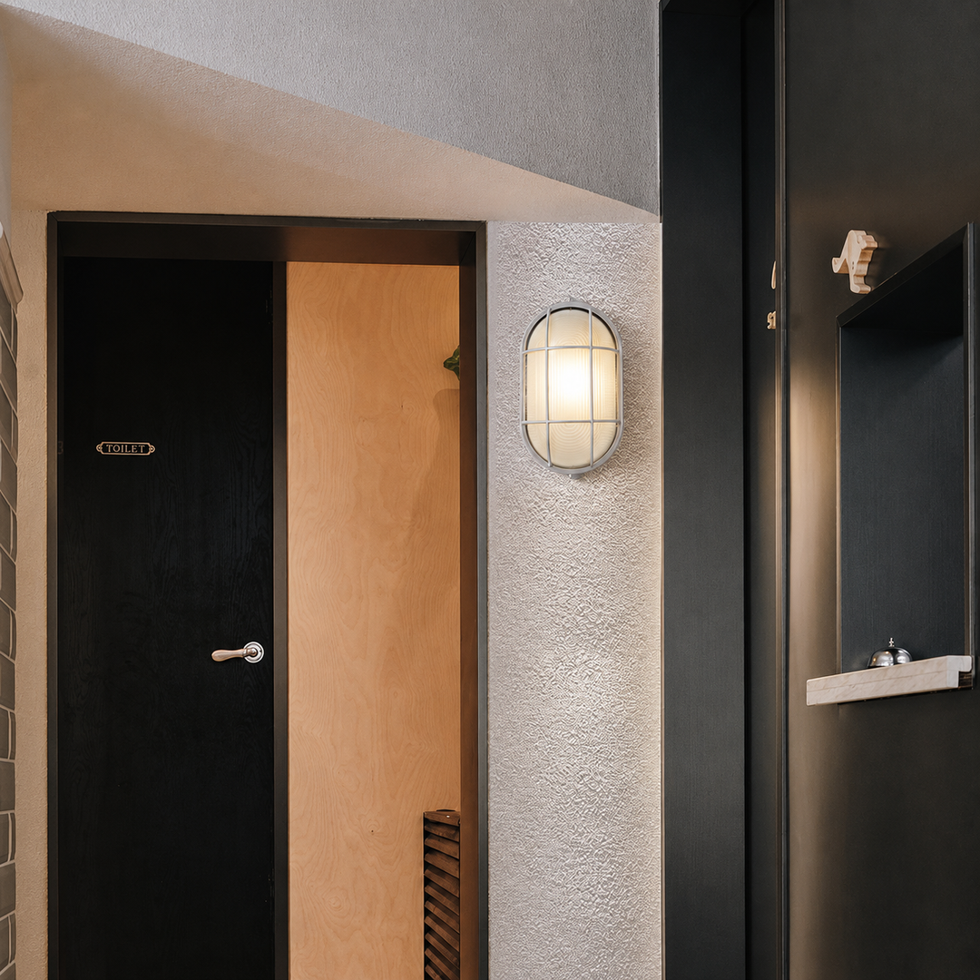 A modern oval wall sconce with a wire grid shade and warm glowing light, mounted on a textured white stucco pillar between two dark doors.