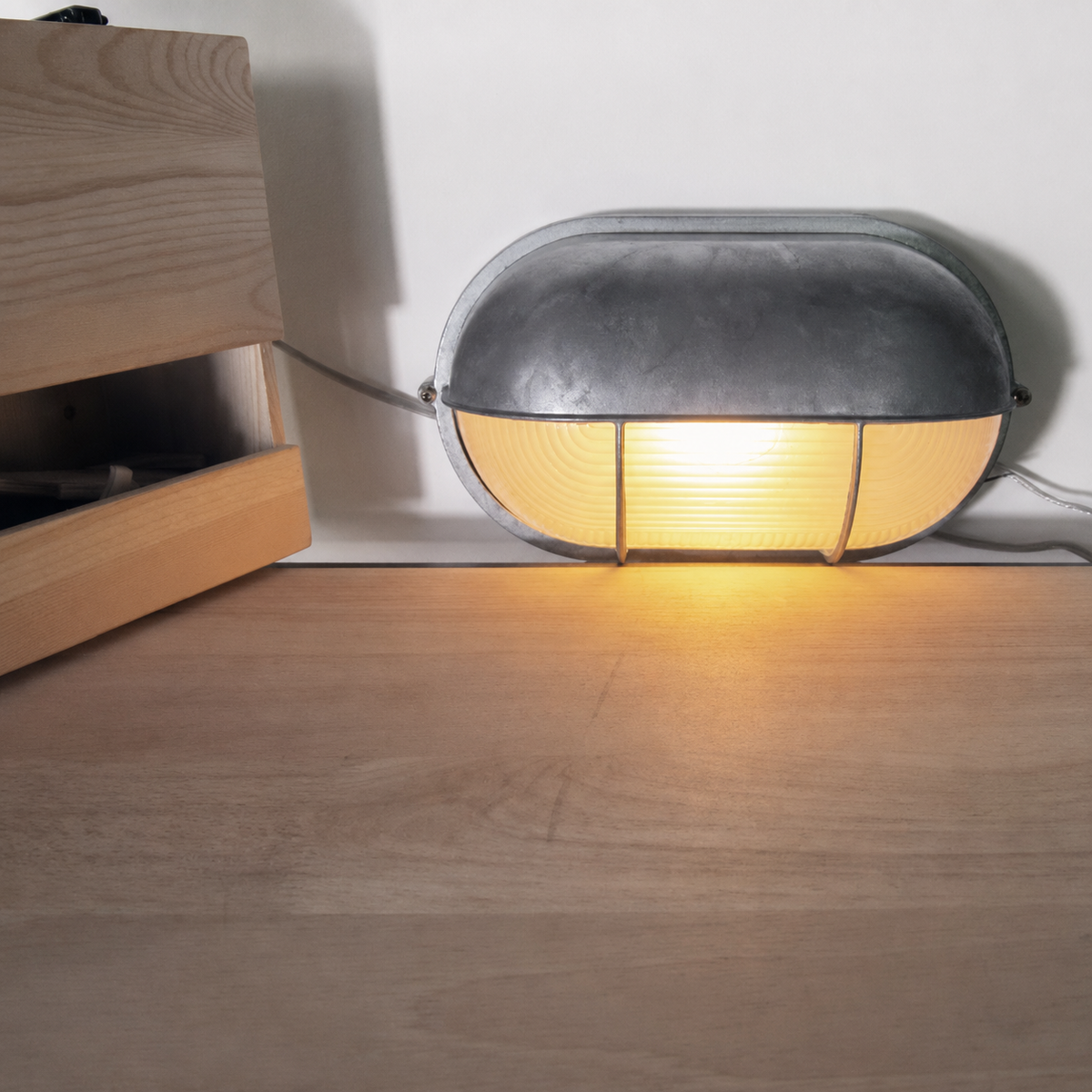 Silver metal wall sconce glowing warm yellow through ribbed glass, resting on a wooden surface beside a wooden crate.