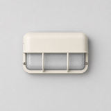 Front view of an unlit off-white wall sconce with ribbed glass, two protective bars, and visible screws against a gray wall.