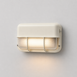 Off-white wall sconce with ribbed glass and protective bars, viewed at an angle and casting warm light down a pale gray wall.