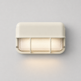 Front view of an illuminated off-white wall sconce with ribbed glass and protective bars, casting warm light onto a gray wall.