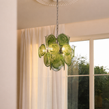 A green glass chandelier with multiple circular shades hangs from a chain in a bright room with white curtains and a window showing outdoor greenery.