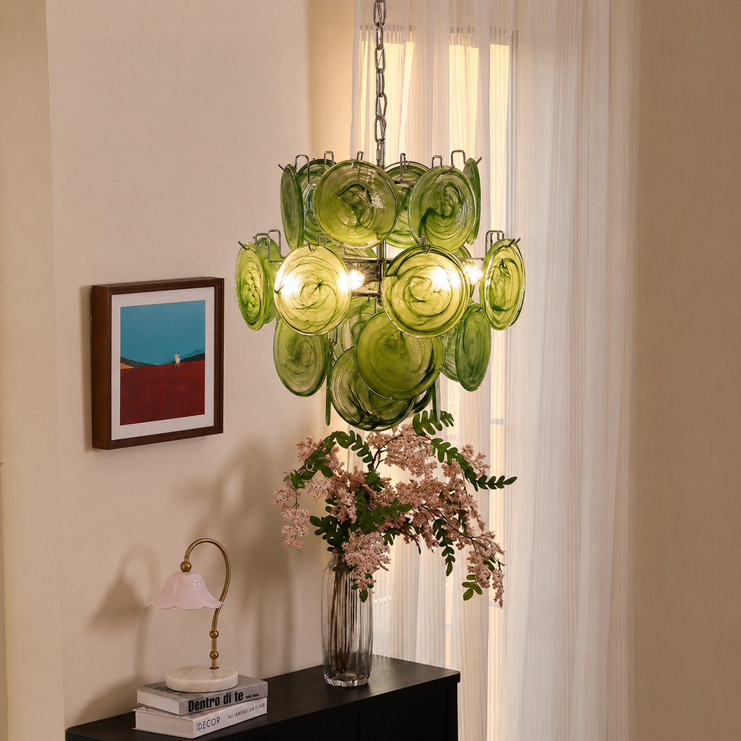 A green glass chandelier with multiple circular shades hangs in a softly lit room with cream curtains, a framed abstract print on the wall, and a black console table holding a vase of pink flowers and a small lamp.