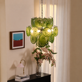 A green glass chandelier with multiple circular shades hangs in a softly lit room with cream curtains, a framed abstract print on the wall, and a black console table holding a vase of pink flowers and a small lamp.