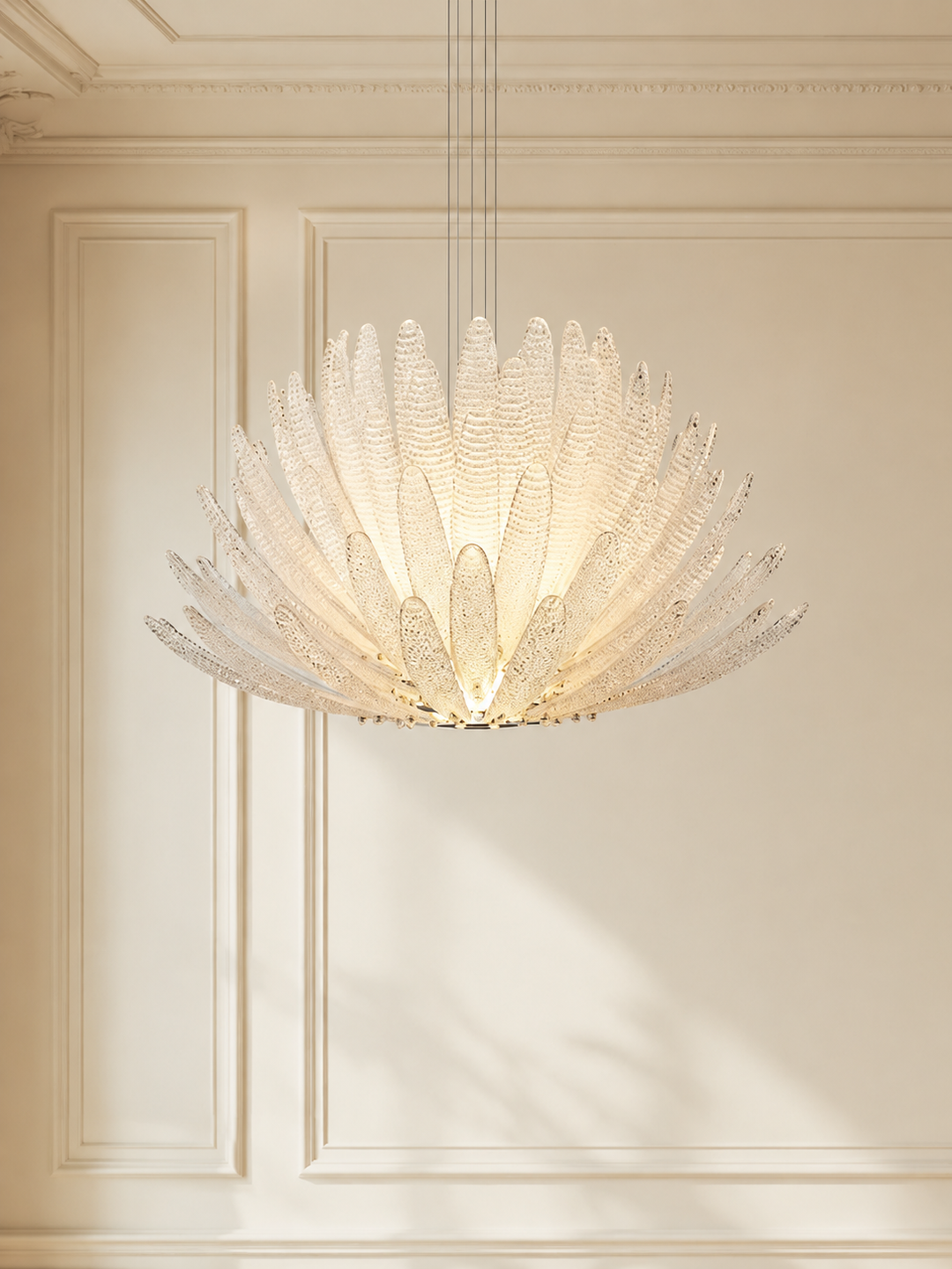 A large, modern flower-shaped chandelier with textured, translucent white glass petals, suspended from thin cables in an elegant room with cream-colored wall paneling.