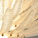 A close-up of a modern chandelier with textured, ribbed glass leaf-like arms glowing with warm light, accented by small gold metal finials.