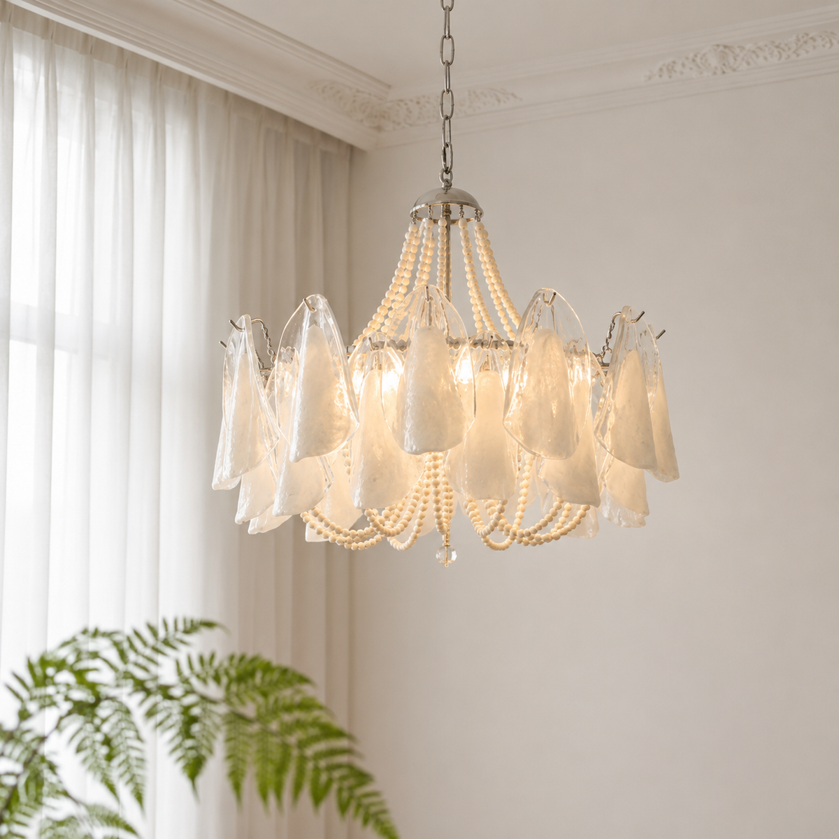 A multi-light chandelier with translucent white bell-shaped glass shades, draped beaded strands, and a silver-toned metal frame, hanging in a bright room with white curtains and a green plant in the foreground.