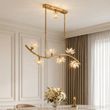 A modern multi-light linear chandelier with a gold-finished frame and clear crystal flower-shaped shades, hanging in a decorated dining room with beige walls, curtains, and a table setting.