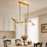 A modern brass six-light linear chandelier with clear glass crystal flower shades hanging over a dining table in a beautifully decorated dining room.