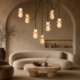 A modern living room scene with a curved beige sofa, round stone coffee table, and a hanging chandelier with multiple organic-shaped glowing light bulbs suspended from black chains against a warm beige wall with an arched alcove.