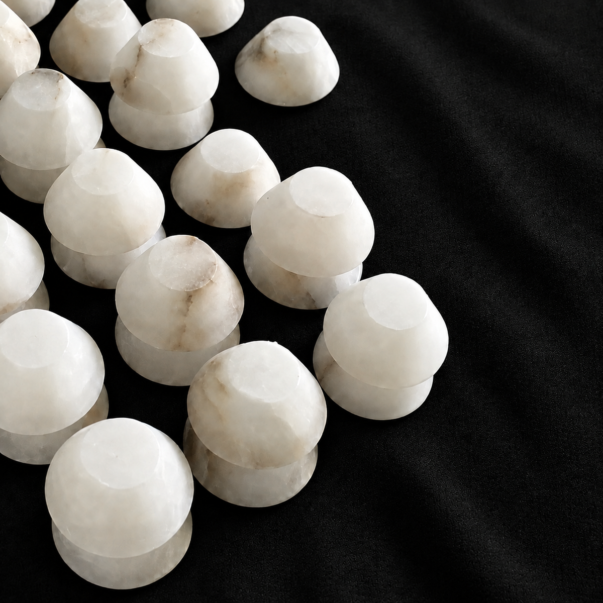 White marble chandelier shade components with subtle beige veining are arranged in rows on textured black fabric.