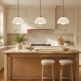 A bright, modern kitchen island with a light wood countertop and two white bar stools, surrounded by cream cabinetry, three hanging pendant lights, and a stone backsplash.