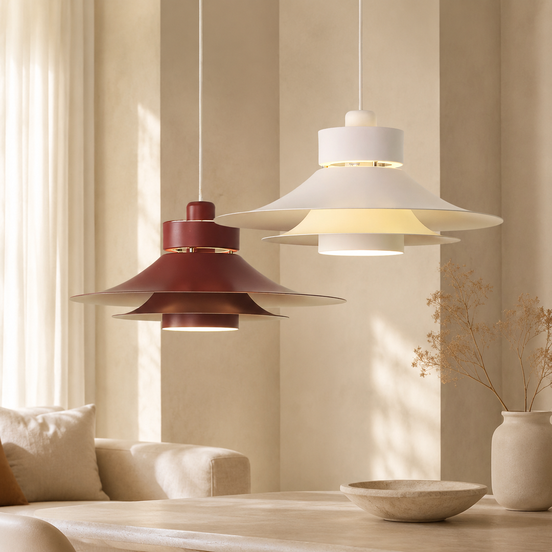 Two modern pendant light fixtures hanging in a softly lit interior. One has a white shade and the other a muted red-brown shade, both with layered circular petals and warm glowing bulbs.