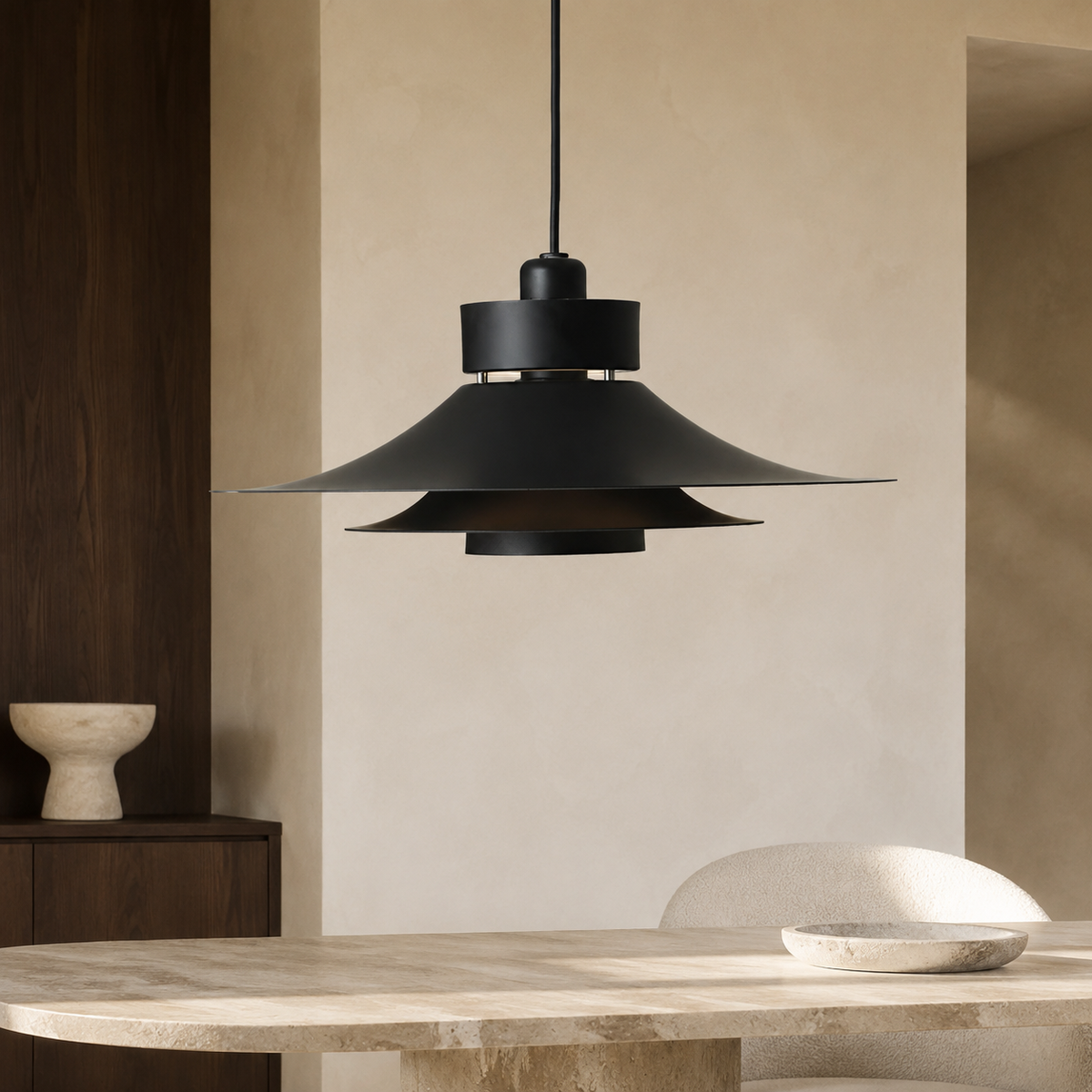A black metal pendant light with a wide, layered conical shade hanging over a light stone dining table in a minimalist, warm-toned interior.